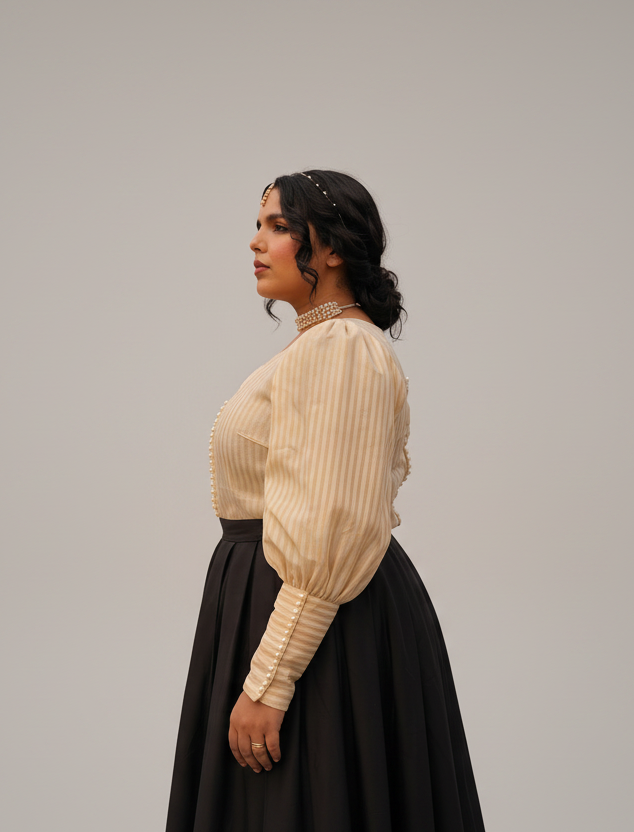 A woman stands in profile in a dry grassy field with palm trees under an overcast sky, wearing a cream ribbed blouse with puff sleeves and a black skirt.