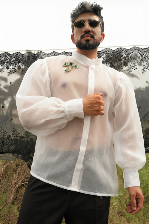 A man wearing a sheer white long-sleeve button-up shirt with a high collar, paired with black trousers and sunglasses, photographed from an overhead angle against a patterned backdrop.