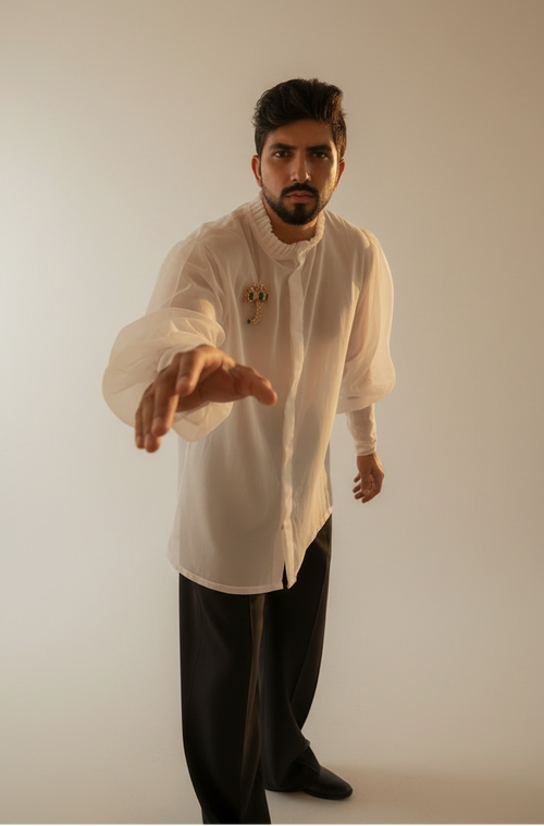 A man wearing a white long-sleeve button-up shirt with a high collar and black trousers, standing outdoors in a grassy field with palm trees under a cloudy sky.