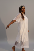 A woman stands outdoors in a grassy field at sunset wearing a loose, semi-sheer white tunic dress with wide sleeves and barefoot. She is adorned with a silver headpiece, necklace, and earrings.